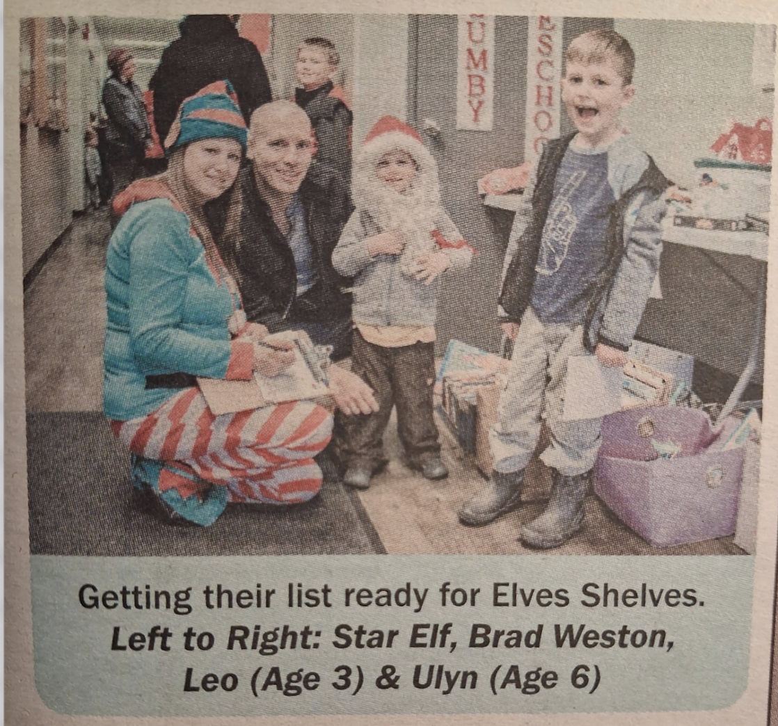Newspaper clipping captioned 'Getting their list ready for Elves Shelves', showing Bradley Weston with an Elves Shelves volunteer and two children in December 2018.