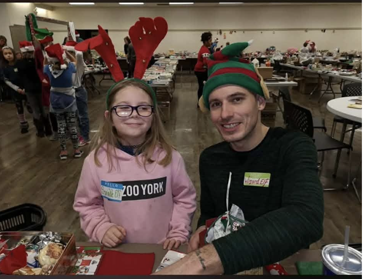 Bradley Weston wearing a green elf hat while volunteering at Elves Shelves in December 2019, with a young helper.